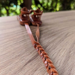 May include: A pair of brown leather sandals with braided straps. The sandals are tied together and laid out on a wooden surface.