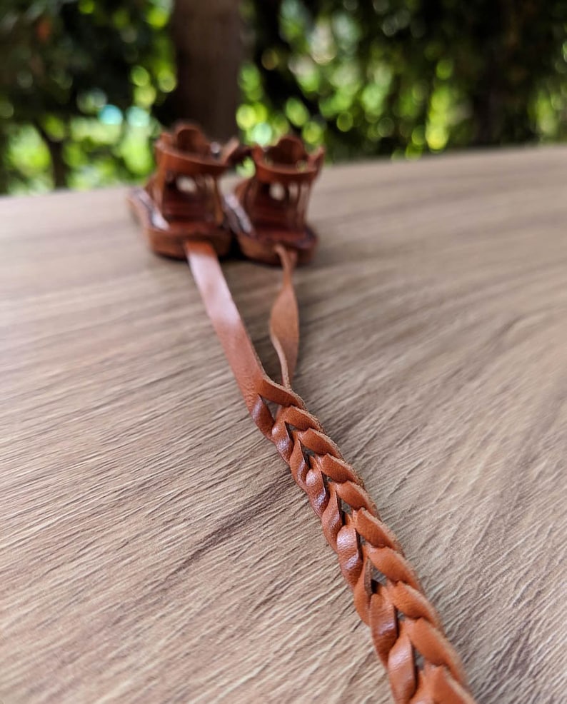 May include: A pair of brown leather sandals with braided straps. The sandals are tied together and laid out on a wooden surface.