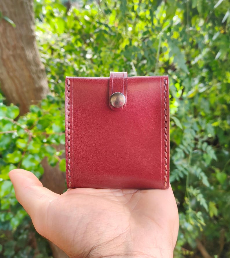 Peut inclure: Un petit portefeuille en cuir bordeaux avec une fermeture &agrave; bouton pression. Le portefeuille a un bord cousu et est tenu dans une main.