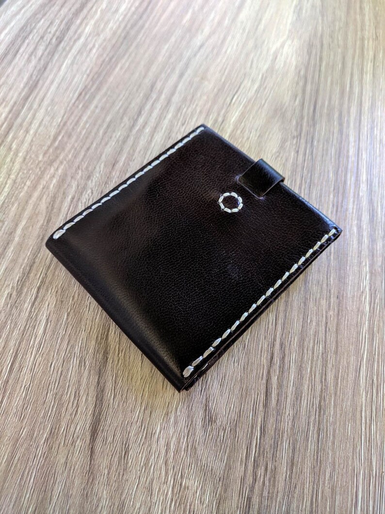 Peut inclure: Un portefeuille en cuir marron fonc&eacute; avec des coutures blanches sur les bords. Le portefeuille a un petit rabat avec une fermeture &agrave; bouton.