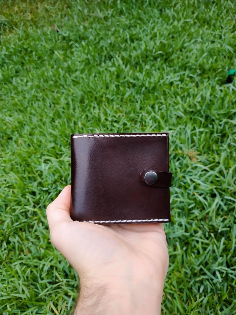 Peut inclure: Un portefeuille en cuir marron avec des coutures blanches et une fermeture &agrave; pression argent&eacute;e.