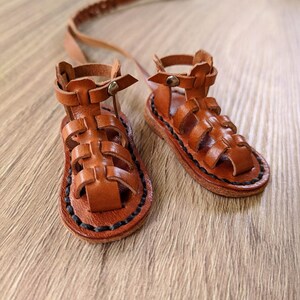 May include: A pair of brown leather sandals with a gladiator style design. The sandals have a black stitching detail around the edges and a strap that goes around the ankle.
