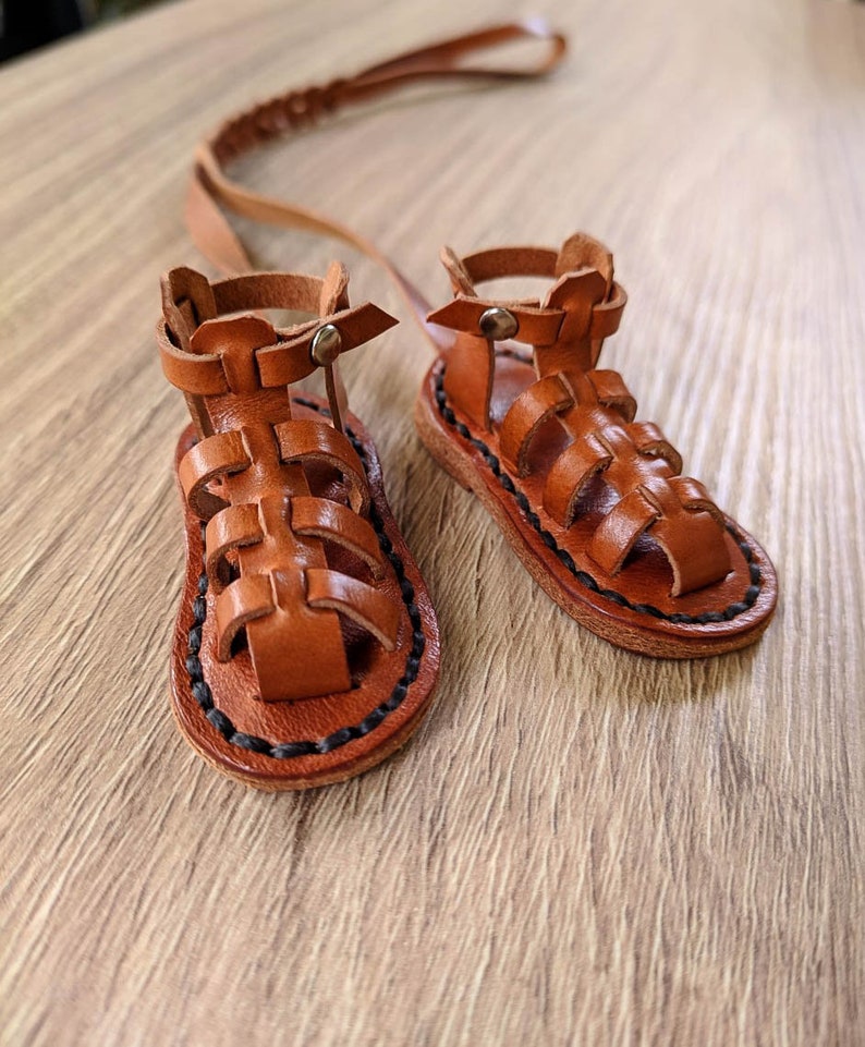 May include: A pair of brown leather sandals with a gladiator style design. The sandals have a black stitching detail around the edges and a strap that goes around the ankle.