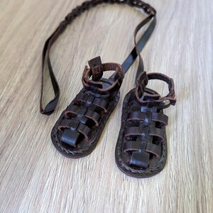 May include: A pair of brown leather sandals with a braided strap. The sandals have a criss-cross design and are tied together with a long leather strap.