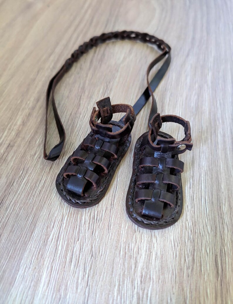 May include: A pair of brown leather sandals with a braided strap. The sandals have a criss-cross design and are tied together with a long leather strap.