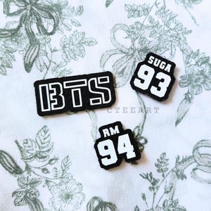 May include: Three black and white patches with the text "BTS", "SUGA 93", and "RM 94". The patches are rectangular and have a white border. The background is a white fabric with a floral pattern.