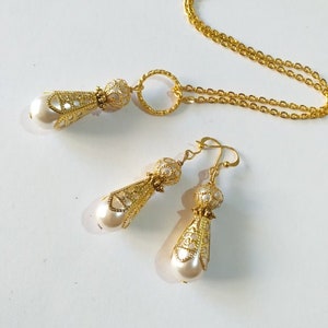 May include: A gold necklace and earrings set with a delicate, ornate design and pearl accents. The necklace features a circular pendant and a chain with a delicate link pattern. The earrings are drop earrings with a similar design to the pendant.