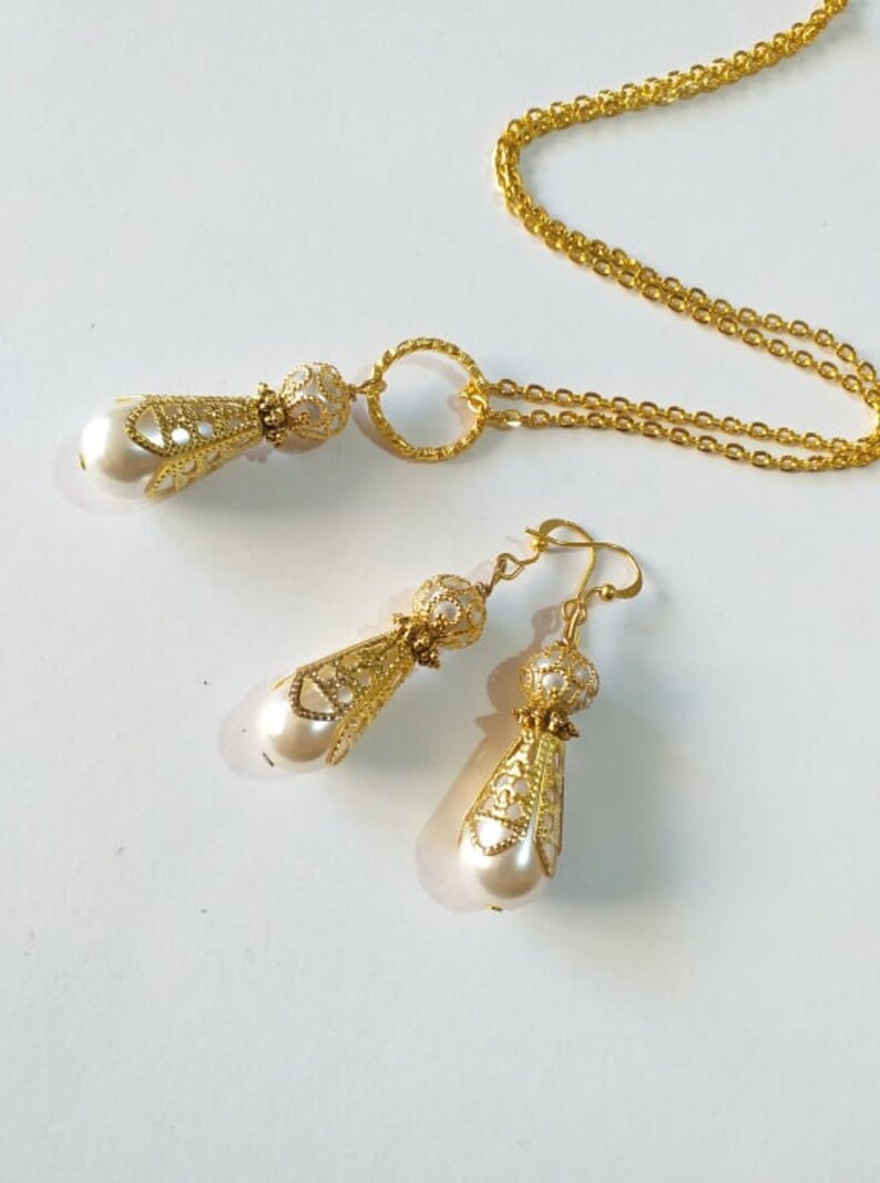 May include: A gold necklace and earrings set with a delicate, ornate design and pearl accents. The necklace features a circular pendant and a chain with a delicate link pattern. The earrings are drop earrings with a similar design to the pendant.