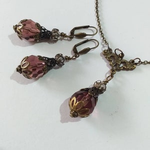 Op de afbeelding: Een set met paarse glazen en bronzen oorbellen en een bijpassende ketting. De oorbellen en ketting hebben een traanvormige paarse glazen kraal met een bronzen filigraanontwerp.