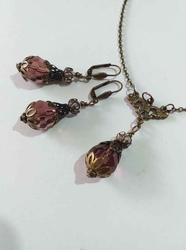 Op de afbeelding: Een set met paarse glazen en bronzen oorbellen en een bijpassende ketting. De oorbellen en ketting hebben een traanvormige paarse glazen kraal met een bronzen filigraanontwerp.