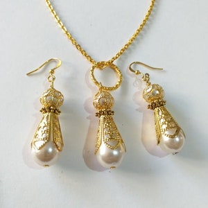 May include: A gold necklace with a circular pendant and matching earrings. The pendant and earrings feature a delicate filigree design and a white pearl drop.