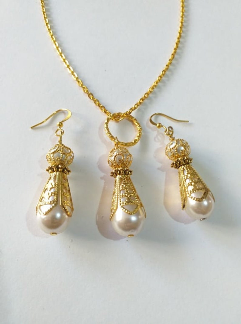 May include: A gold necklace with a circular pendant and matching earrings. The pendant and earrings feature a delicate filigree design and a white pearl drop.