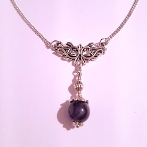 May include: A silver chain necklace with a silver pendant featuring a floral design. The pendant is attached to a single, round, dark purple gemstone.