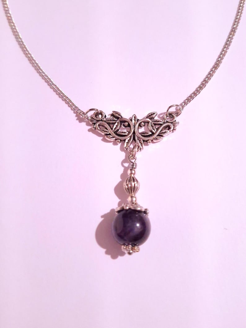 May include: A silver chain necklace with a silver pendant featuring a floral design. The pendant is attached to a single, round, dark purple gemstone.