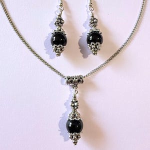 May include: A jewelry set featuring a necklace and matching earrings. The set includes a silver-toned chain necklace with a pendant and two dangle earrings. Each piece has a large, round, black bead accented with silver-colored filigree and decorative elements.