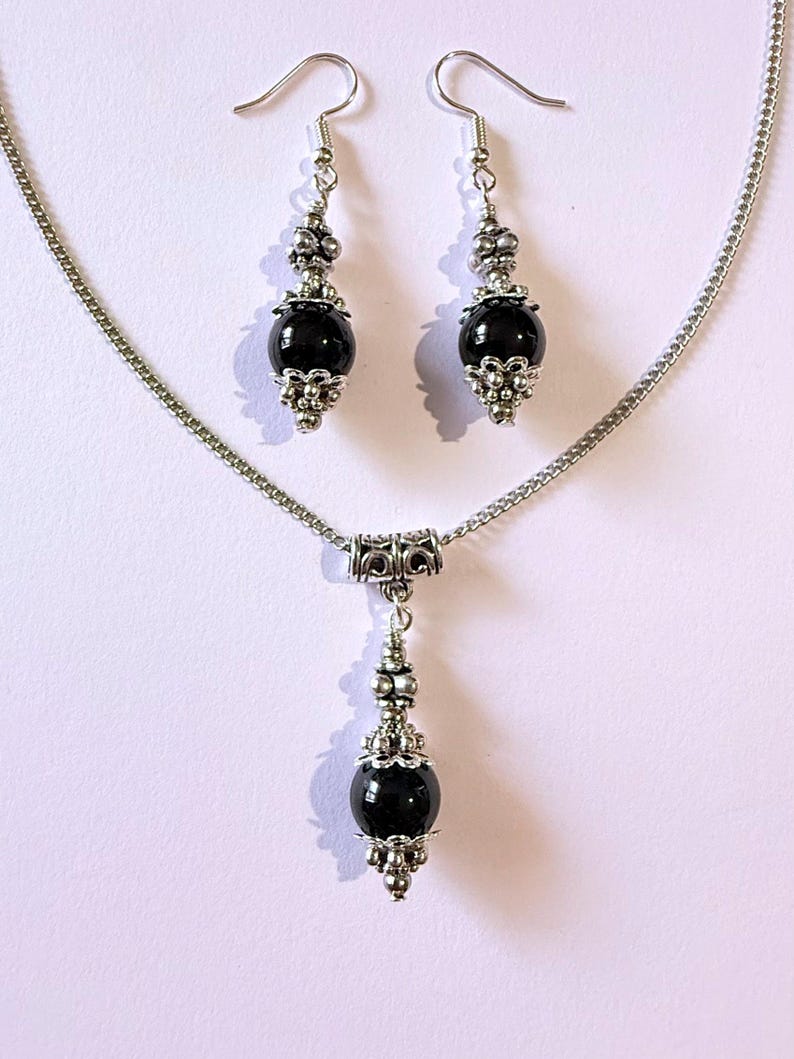 May include: A jewelry set featuring a necklace and matching earrings. The set includes a silver-toned chain necklace with a pendant and two dangle earrings. Each piece has a large, round, black bead accented with silver-colored filigree and decorative elements.