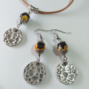 May include: A jewelry set featuring a necklace and earrings. The necklace has a brown leather cord with a pendant of a yellow and black bead and a silver hammered disc. The earrings have matching beads and discs.