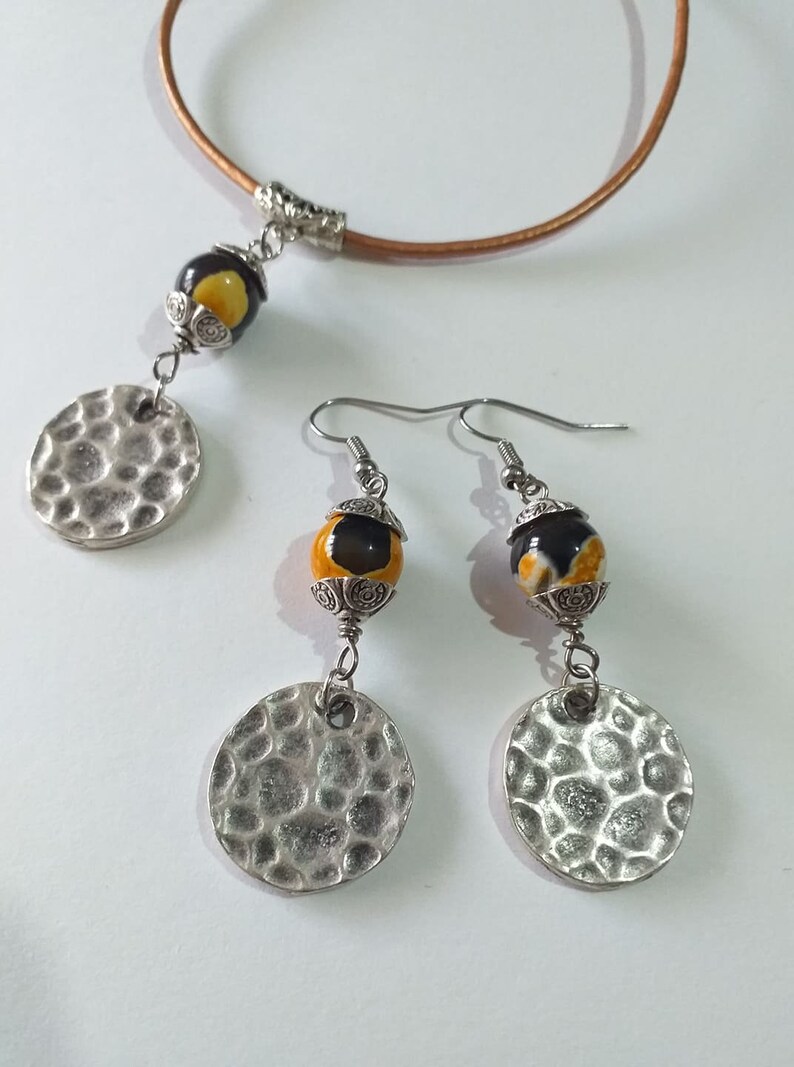 May include: A jewelry set featuring a necklace and earrings. The necklace has a brown leather cord with a pendant of a yellow and black bead and a silver hammered disc. The earrings have matching beads and discs.