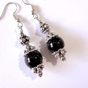 May include: A pair of dangle earrings featuring large, glossy black beads. The earrings have silver-colored metal accents, including small beads and decorative filigree. The hooks are also silver-colored, creating a striking contrast with the dark beads.