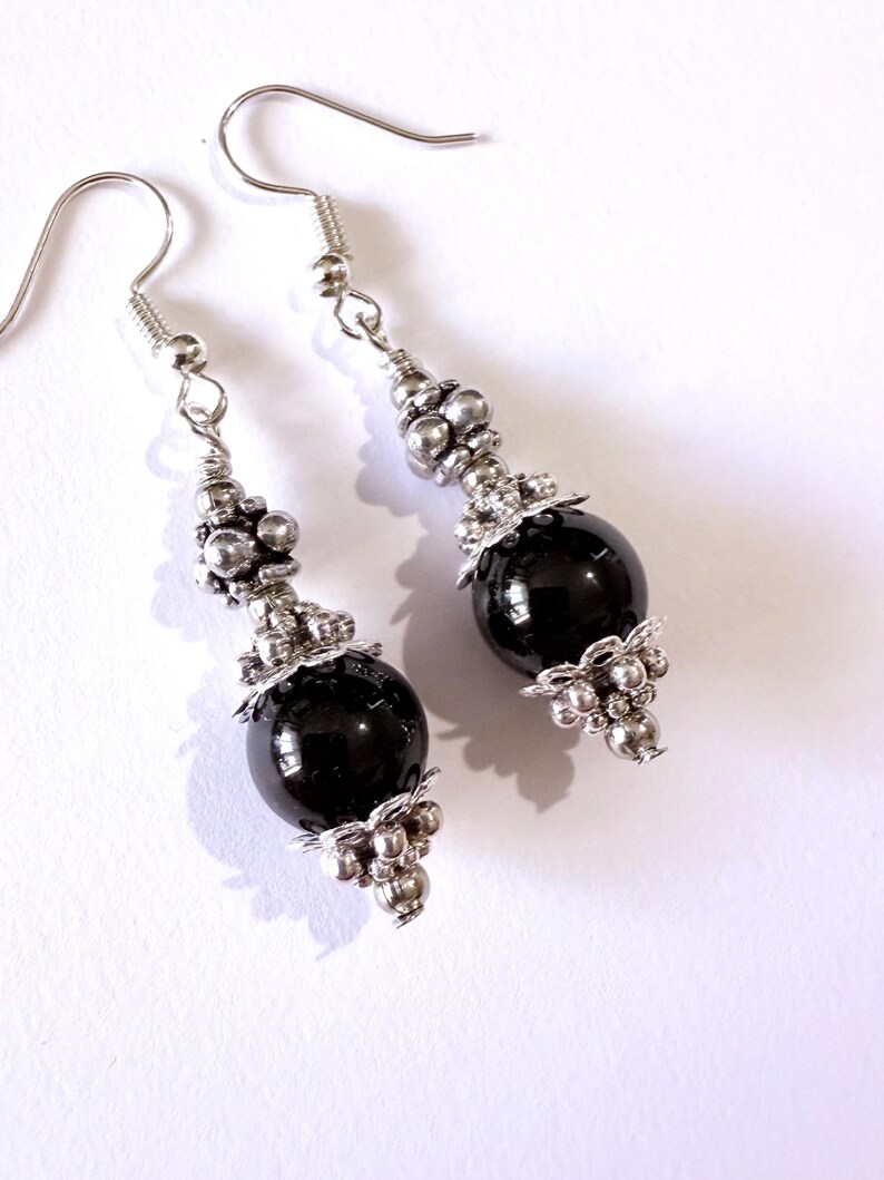 May include: A pair of dangle earrings featuring large, glossy black beads. The earrings have silver-colored metal accents, including small beads and decorative filigree. The hooks are also silver-colored, creating a striking contrast with the dark beads.