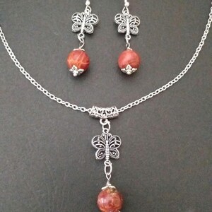 May include: A silver chain necklace with a red stone pendant and a silver butterfly charm. Matching earrings with red stones and butterfly charms.