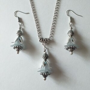 May include: A silver necklace and earrings set featuring a delicate flower pendant and matching earrings. The flower is made from a light blue material and has a silver bead at its centre.