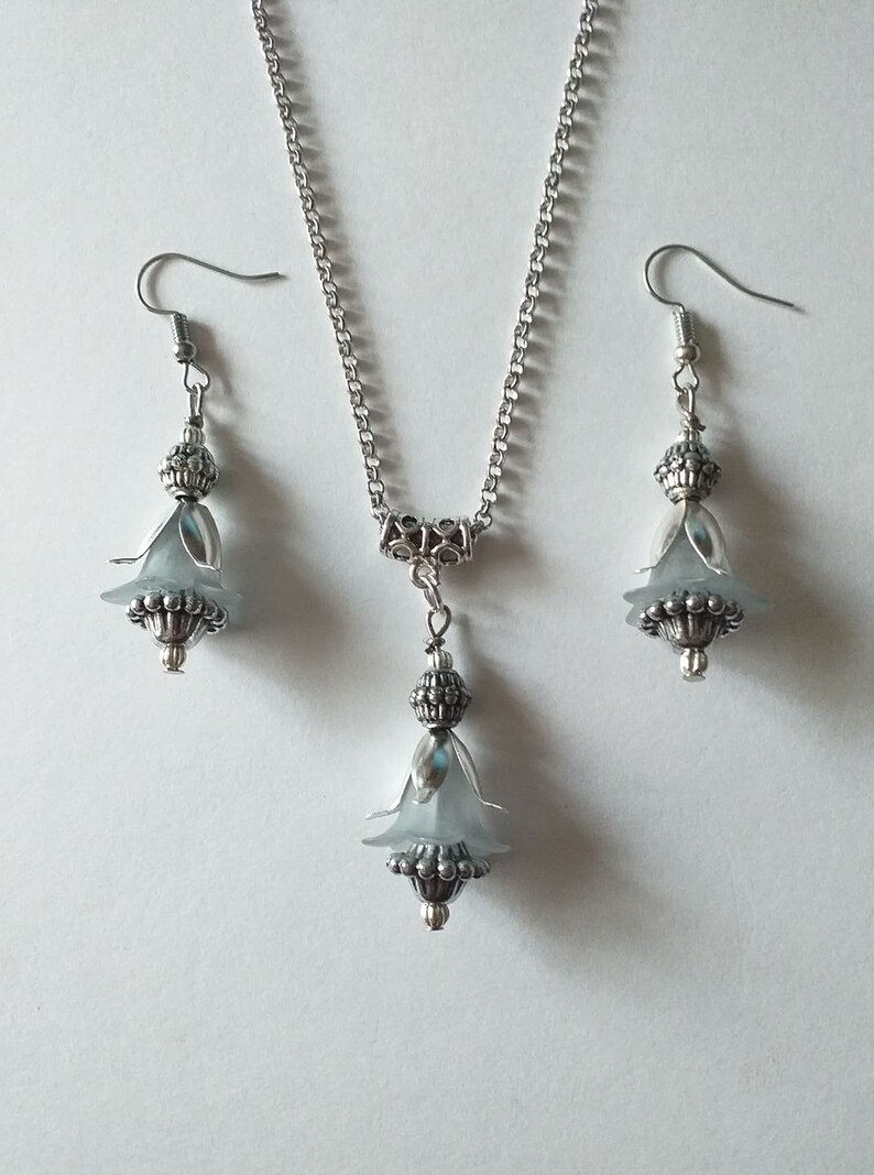 May include: A silver necklace and earrings set featuring a delicate flower pendant and matching earrings. The flower is made from a light blue material and has a silver bead at its centre.