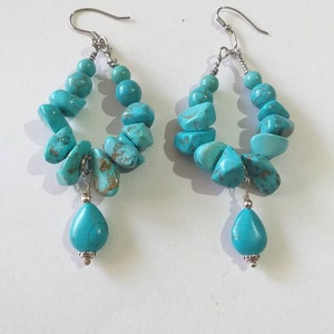 May include: A pair of turquoise dangle earrings with a teardrop shaped turquoise stone hanging from each hoop. The hoops are made of silver and are decorated with turquoise stones.
