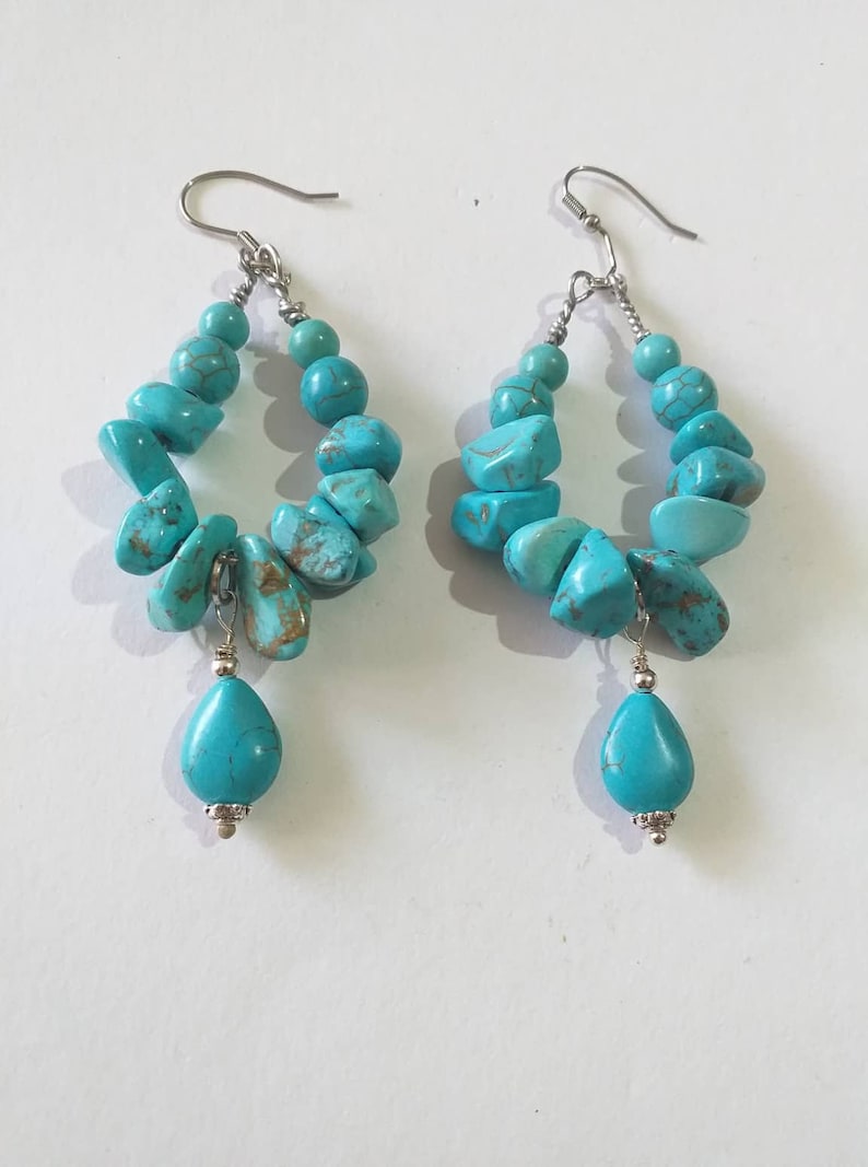 May include: A pair of turquoise dangle earrings with a teardrop shaped turquoise stone hanging from each hoop. The hoops are made of silver and are decorated with turquoise stones.