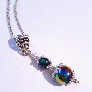 Op de afbeelding: Een zilveren ketting met een zilveren bedel, een groene kraal en een grote, iriserende kraal.