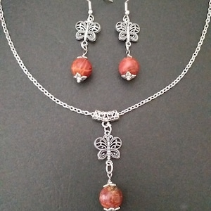 May include: A silver chain necklace with a red and brown stone pendant and a silver butterfly charm. Matching earrings with red and brown stone drops and silver butterfly charms.