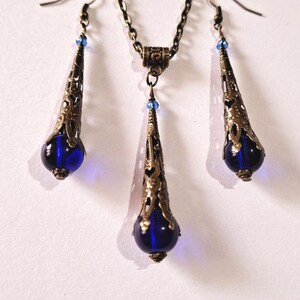 May include: A set of blue glass and bronze jewelry featuring a necklace and earrings. The necklace and earrings have a teardrop shape with a blue glass bead at the bottom. The bronze is an antique gold color.