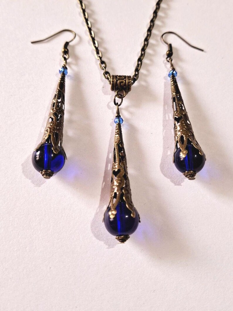 May include: A set of blue glass and bronze jewelry featuring a necklace and earrings. The necklace and earrings have a teardrop shape with a blue glass bead at the bottom. The bronze is an antique gold color.