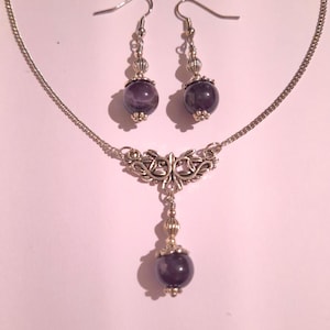 May include: A silver necklace with a pendant featuring a silver filigree design and a purple amethyst stone. Matching earrings with silver hooks and purple amethyst stones.