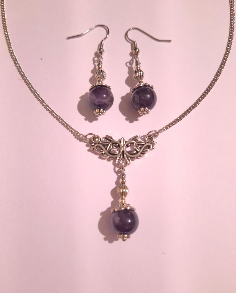 May include: A silver necklace with a pendant featuring a silver filigree design and a purple amethyst stone. Matching earrings with silver hooks and purple amethyst stones.