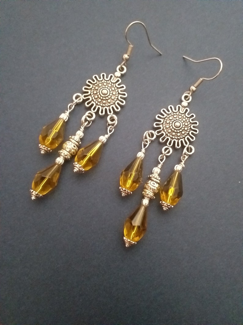 May include: A pair of silver dangle earrings with a round, ornate design at the top and three amber-colored teardrop-shaped beads hanging from each.