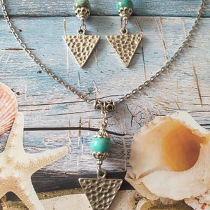 May include: A silver necklace and earrings set with turquoise stones and triangle pendants. The necklace has a silver chain and a turquoise stone with a silver pendant in the shape of a triangle. The earrings have silver hooks and turquoise stones with silver triangle pendants.