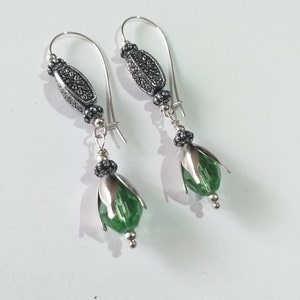 May include: A pair of silver earrings with green crystal drops. The earrings feature a silver loop with a decorative bead and a smaller bead below it. The crystal drops are faceted and have a green hue.