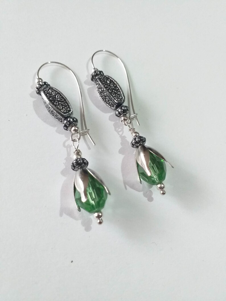May include: A pair of silver earrings with green crystal drops. The earrings feature a silver loop with a decorative bead and a smaller bead below it. The crystal drops are faceted and have a green hue.