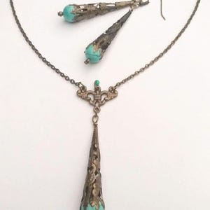 Puede incluir: Un conjunto de collar y pendientes de turquesa y bronce. El collar presenta una cadena delicada con un colgante que tiene una piedra turquesa engastada en un dise&ntilde;o de filigrana de bronce. Los pendientes son pendientes colgantes con una piedra turquesa y un dise&ntilde;o de filigrana de bronce similares.