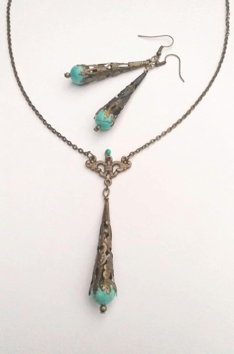 Puede incluir: Un conjunto de collar y pendientes de turquesa y bronce. El collar presenta una cadena delicada con un colgante que tiene una piedra turquesa engastada en un dise&ntilde;o de filigrana de bronce. Los pendientes son pendientes colgantes con una piedra turquesa y un dise&ntilde;o de filigrana de bronce similares.
