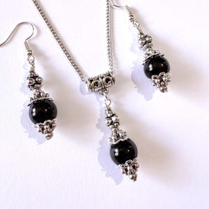 May include: A silver-toned jewelry set featuring a necklace and matching earrings. The necklace has a chain with a pendant, and the earrings dangle. Each piece has a black, round bead accented with silver-colored metalwork.