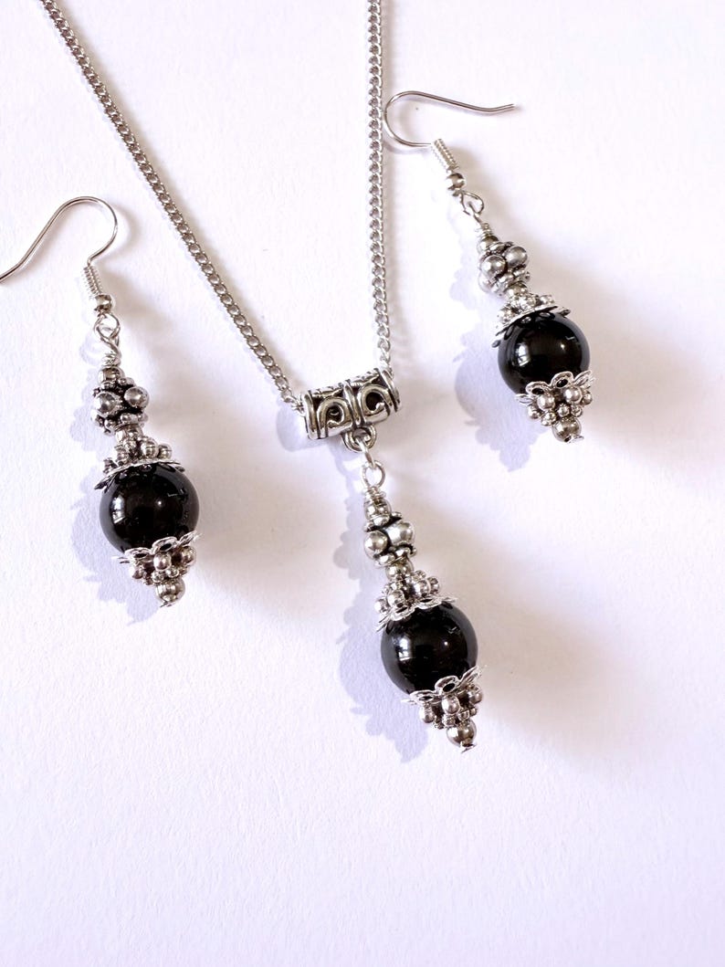 May include: A silver-toned jewelry set featuring a necklace and matching earrings. The necklace has a chain with a pendant, and the earrings dangle. Each piece has a black, round bead accented with silver-colored metalwork.