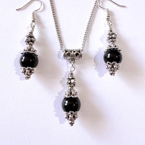 May include: A silver necklace and earring set featuring black, spherical beads. The necklace has a decorative pendant, and the earrings have a similar design. The jewelry set is displayed on a white background.