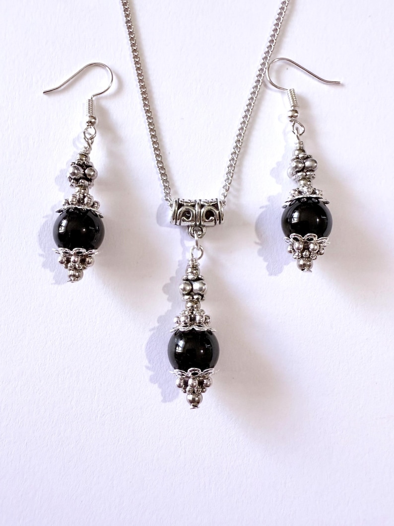 May include: A silver necklace and earring set featuring black, spherical beads. The necklace has a decorative pendant, and the earrings have a similar design. The jewelry set is displayed on a white background.