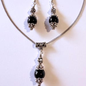 May include: A silver-toned jewelry set featuring a necklace and matching earrings. The necklace has a pendant with a large, black, spherical bead. The earrings also have black beads. Ornate silver-colored metalwork accents the beads.