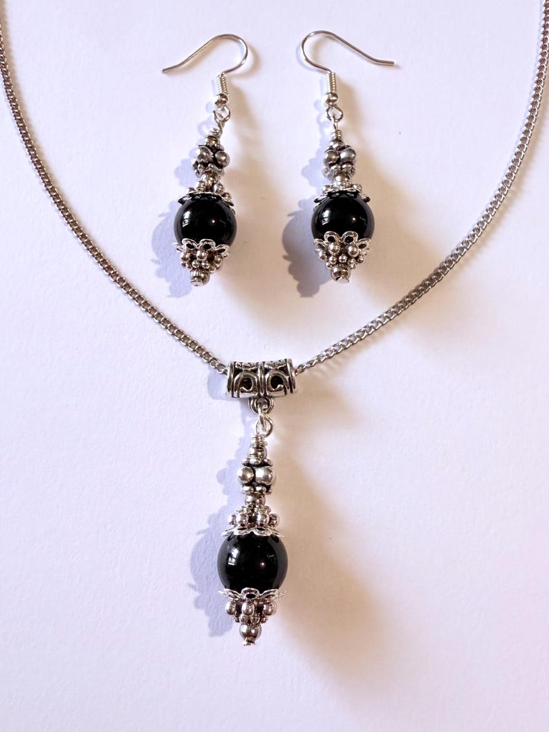 May include: A silver-toned jewelry set featuring a necklace and matching earrings. The necklace has a pendant with a large, black, spherical bead. The earrings also have black beads. Ornate silver-colored metalwork accents the beads.