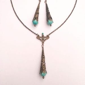 Puede incluir: Un conjunto de collar y pendientes de turquesa y bronce. El collar presenta una cadena delicada con un colgante que tiene una piedra turquesa engastada en un dise&ntilde;o de filigrana. Los pendientes son pendientes colgantes con un dise&ntilde;o similar.