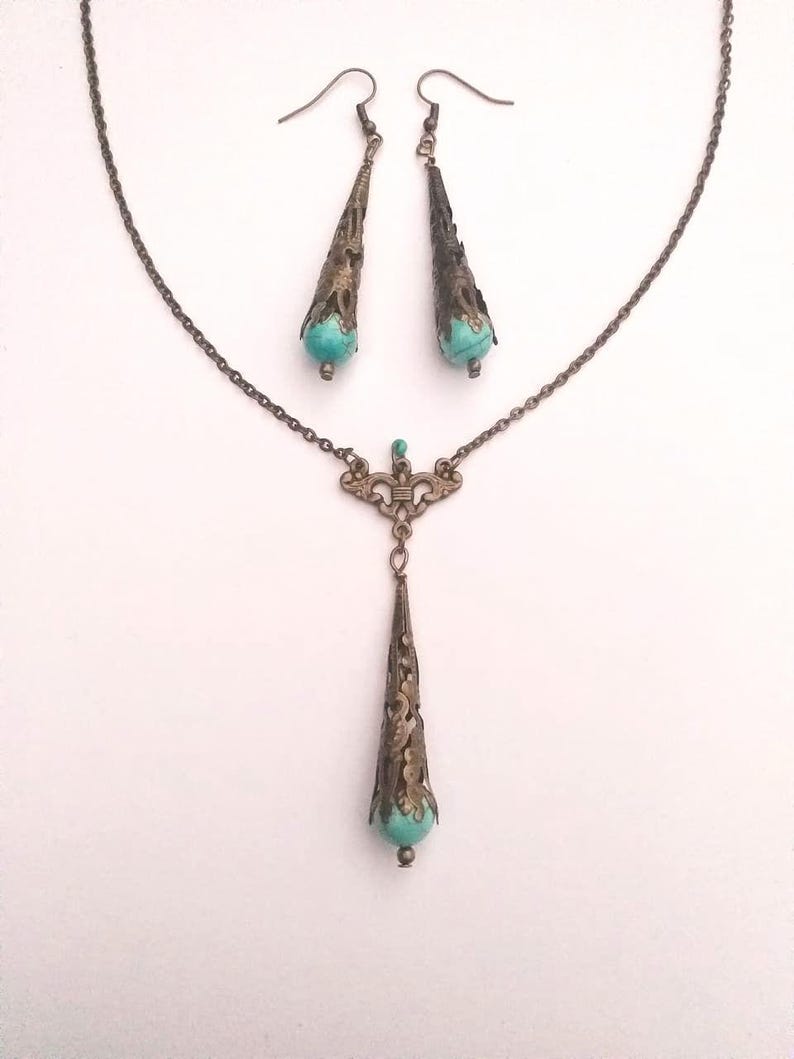 Puede incluir: Un conjunto de collar y pendientes de turquesa y bronce. El collar presenta una cadena delicada con un colgante que tiene una piedra turquesa engastada en un dise&ntilde;o de filigrana. Los pendientes son pendientes colgantes con un dise&ntilde;o similar.