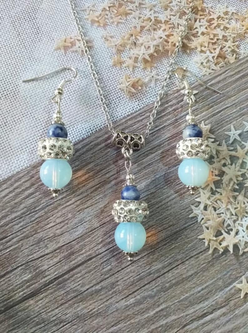 May include: A silver necklace and earrings set with blue and white beads. The necklace has a silver chain and a pendant with a blue and white bead. The earrings have a silver hook and a blue and white bead.